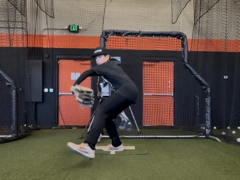 A young pitcher mid-delivery off a portable rubber during an indoor lesson