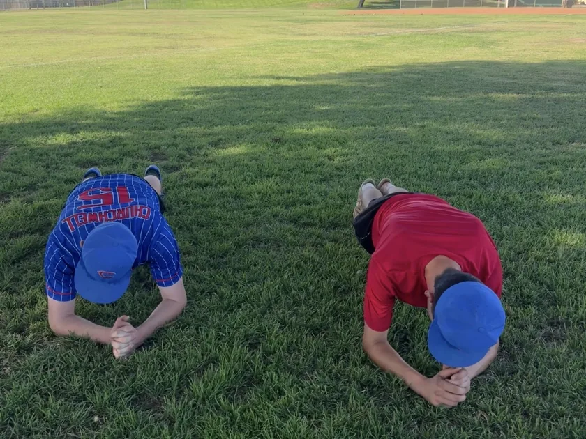 Two young baseball players holding forearm planks on the outfield grass during a training session